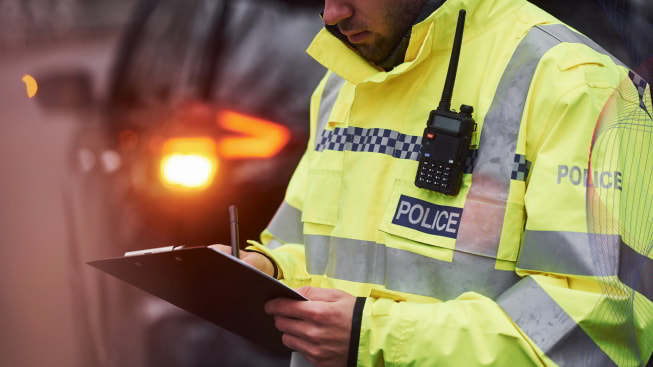 A male police officer in a green uniform standing with a notepad and a radio near a car