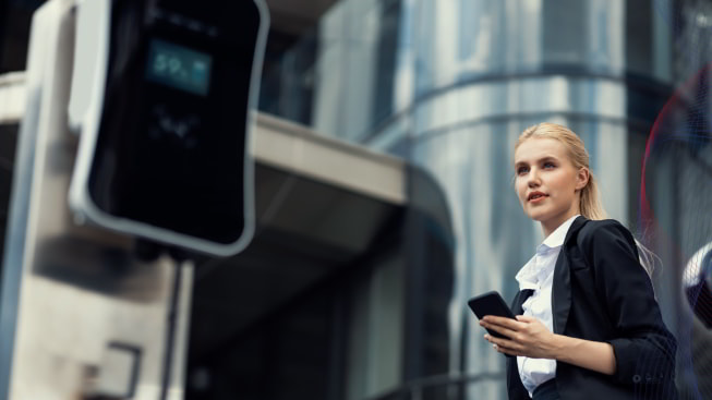 A woman with smartphone at a charging station with an office building in the background